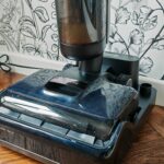 Aspirateur laveur Dreame H15 Pro posé sur sa station, sur un parquet en point de Hongrie, devant un papier peint fleuri