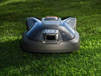 Robot tondeuse Husqvarna Automower sur une pelouse verte, illustrant l'offre d'extension de garantie de 5 ans et la technologie de navigation par vision IA sans câble périphérique.