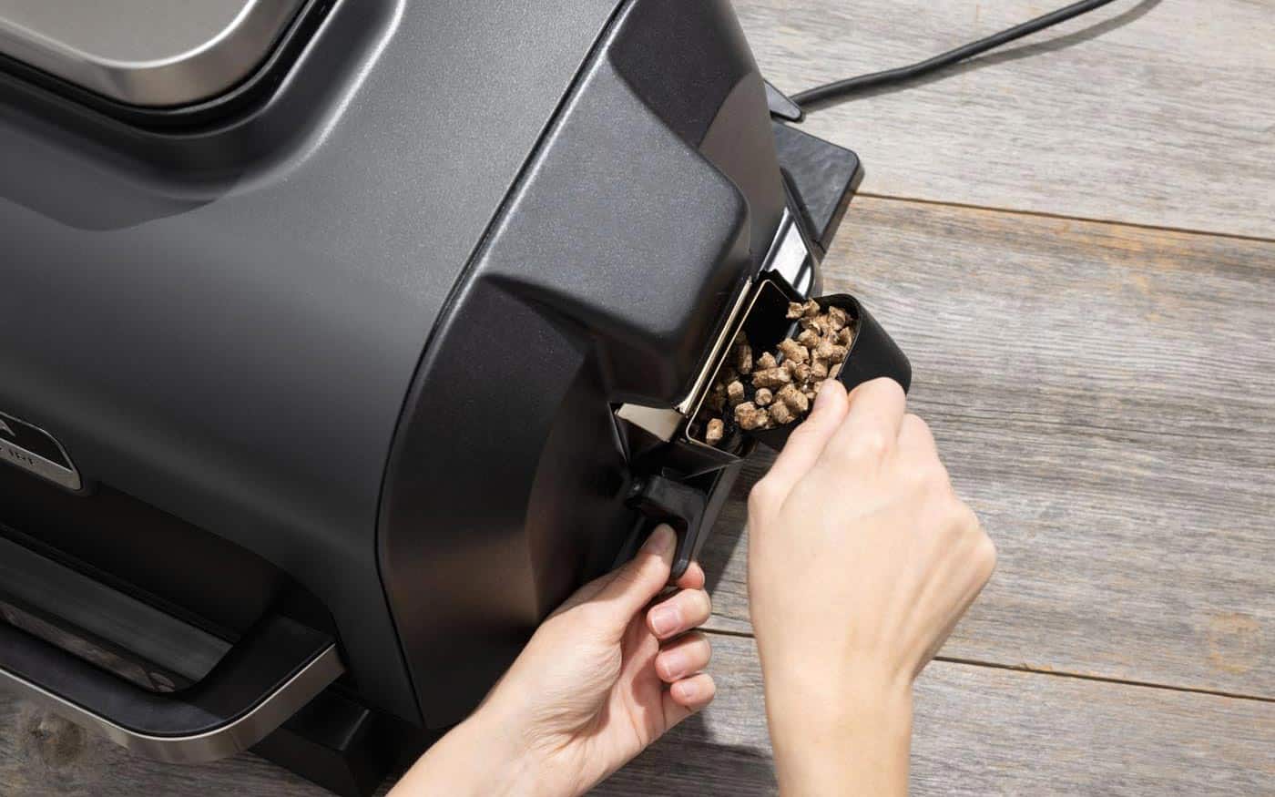 Ajout de granulés de bois naturel dans le bac latéral du fumoir du barbecue électrique Ninja Woodfire Pro XL.