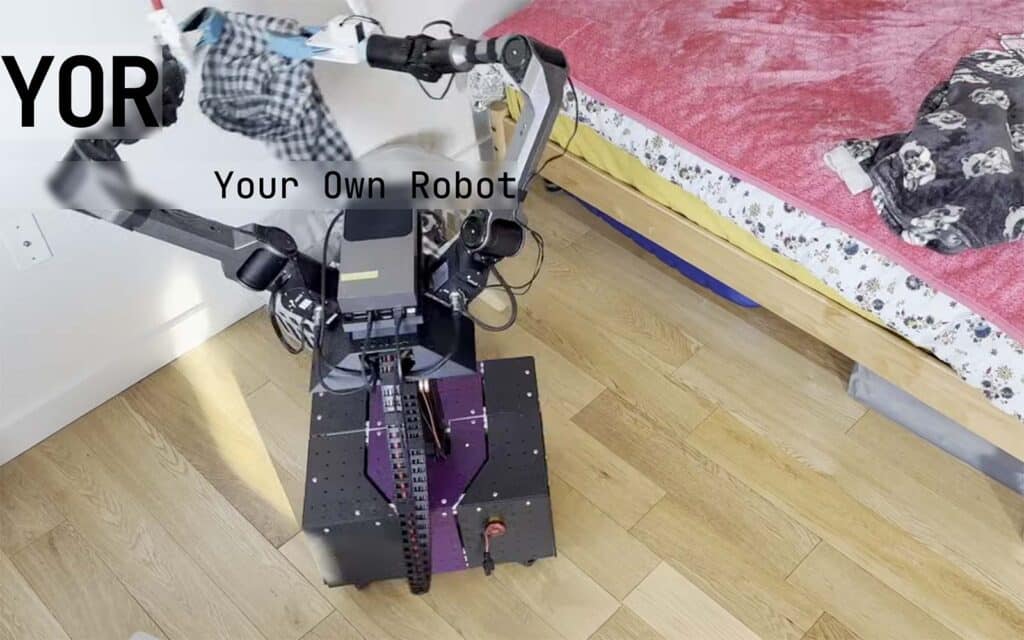 Photo du robot open source YOR dans une chambre à coucher. Le robot utilise ses deux bras articulés mettre du linge dans un panier à linge au sol à côté de lui.