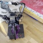 Photo du robot open source YOR dans une chambre à coucher. Le robot utilise ses deux bras articulés mettre du linge dans un panier à linge au sol à côté de lui.