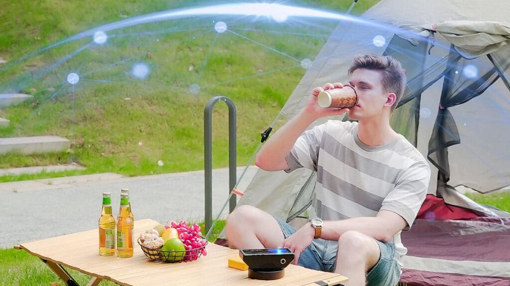 Homme buvant un café en camping à côté d'une tente, protégé des insectes par un dôme virtuel généré par le boîtier anti-moustiques portable Photon Matrix posé sur une table de pique-nique.