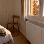 Une main tourne le bouton de réglage d’un radiateur électrique blanc sous la fenêtre d’une chambre d’amis inoccupée, lit fait