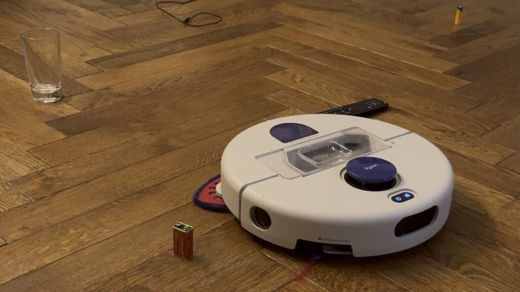 Le robot Dyson R2 Nurovi sur un parquet, entouré d’un verre, d’une pile 9 V, d’une pile AA, d’une télécommande et d’un câble