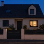 Façade d’une maison de lotissement à la tombée de la nuit, une seule fenêtre éclairée au rez-de-chaussée, les autres fenêtres sombres