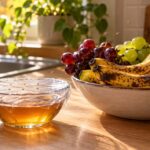 Un bol de vinaigre de cidre couvert d’un film plastique percé, posé sur un plan de travail près d’une coupe de bananes mûres, avec une plante en pot sur le rebord de fenêtre