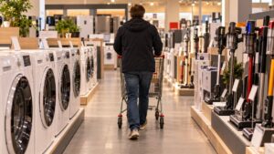 Un client pousse un chariot dans l'allée d'un magasin d'électroménager, entre des lave-linge et des aspirateurs